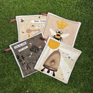 May include: Four brown and beige pot holders with bee-themed designs. The pot holders feature the text "Love Our Bees", "Plant Flowers", "Make Honey, Not War", and "Make a Buzz, Save Our Bees".