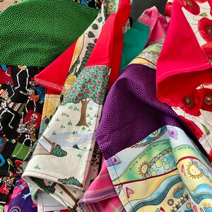 May include: A selection of patterned fabric napkins in a variety of designs. The napkins feature prints such as red poppies, green polka dots, Day of the Dead skeletons, and illustrated scenes. They are folded and layered, displaying the different patterns.