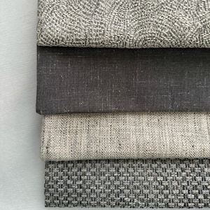 May include: A stack of four fabric swatches in shades of gray and neutral tones. The top swatch has a textured, patterned design. The other swatches have varying textures, including a solid dark gray, a light gray weave, and a darker gray basketweave.