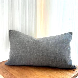 May include: A rectangular gray decorative pillow with a textured fabric. The pillow is placed on a wooden surface, with a white curtain in the background. The pillow's dimensions are not visible.