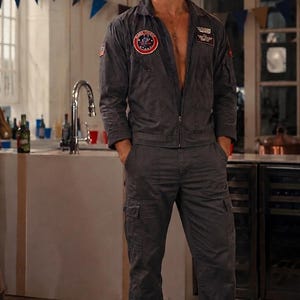May include: A charcoal gray flight suit featuring a front zipper closure, long sleeves, and cargo-style pockets on the legs. The garment includes circular and rectangular embroidered patches on the chest and sleeves, worn with black boots.