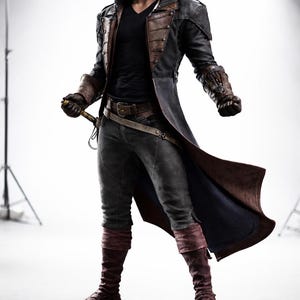May include: A costume featuring a long black coat with brown leather accents, a hooded cloak, and matching gloves. The outfit includes dark grey trousers, burgundy leg warmers, and a belt with a sword. The overall theme is a historical or fantasy character.
