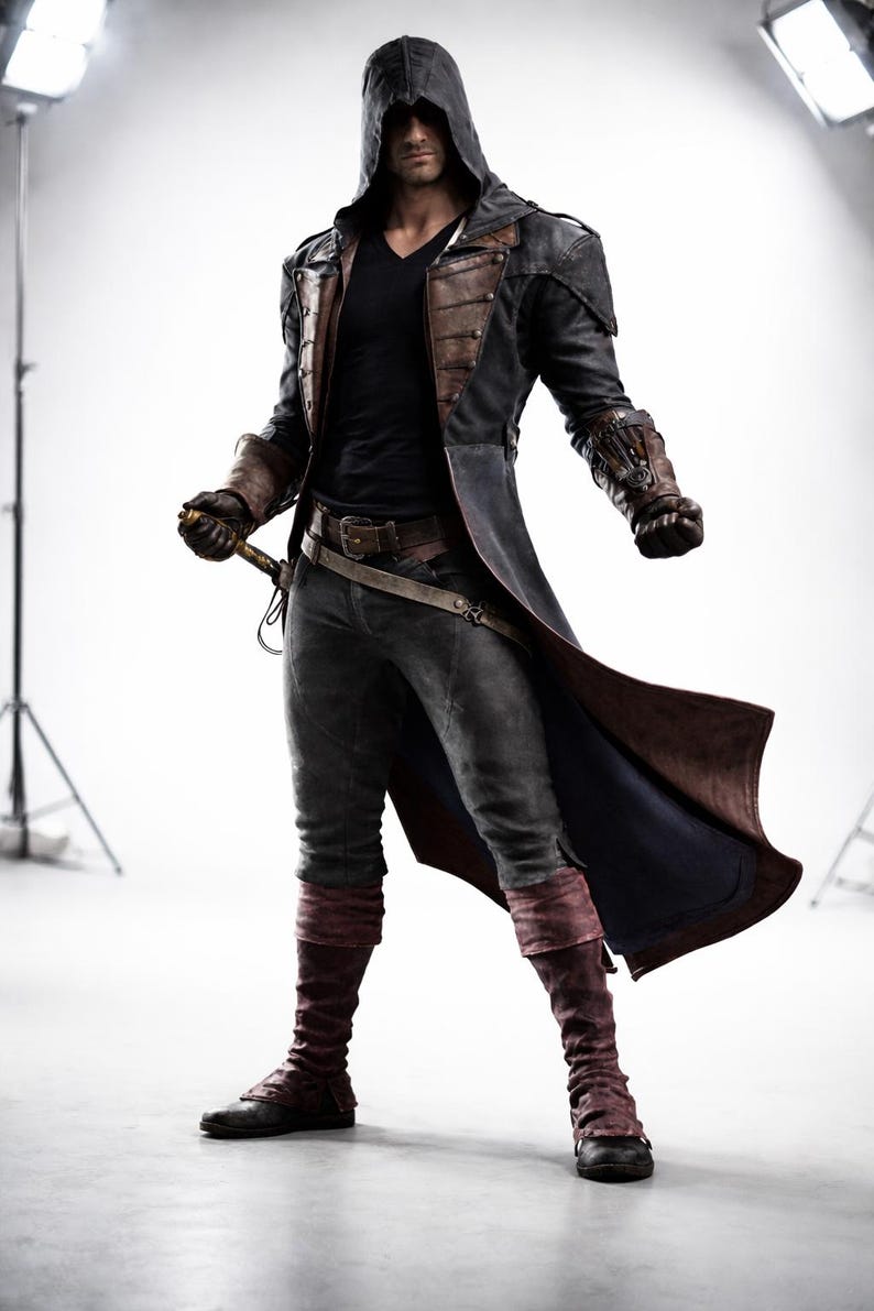 May include: A costume featuring a long black coat with brown leather accents, a hooded cloak, and matching gloves. The outfit includes dark grey trousers, burgundy leg warmers, and a belt with a sword. The overall theme is a historical or fantasy character.