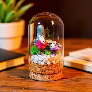 以下が含まれることがあります： 白いと赤色の2つの小さなフィギュアが特徴の装飾的なガラスドームテラリウム。白い石、緑の苔、青い石の構造のミニチュア風景の中に配置されています。テラリウムはコルクベースの上にあります。