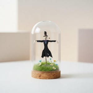 May include: A miniature scarecrow figure in a black suit and top hat, with a pipe, enclosed in a glass dome. The base has green grass, small flowers, and a cork rim. The figure's arms are outstretched.