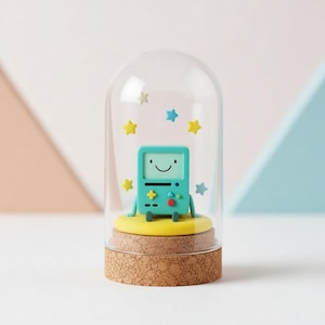 Puede incluir: Figura de personaje turquesa con una cara sonriente, dentro de una cúpula de cristal. La figura está sobre una base amarilla encima de una base de corcho, con decoraciones de estrellas amarillas y azules. El fondo es una mezcla de rosa y azul.