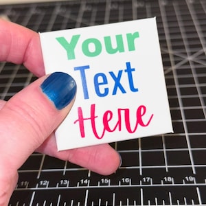 May include: A white square with the words "Your Text Here" in green, blue, and red. The square is held by a hand with blue nail polish. The background is a black cutting mat with white grid lines and numbers.