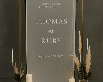 Könnte beinhalten: Ein Hochzeitsschild mit den Namen "Thomas & Ruby" und dem Datum "27. September 2025" auf einem transparenten Stoff. Das Schild wird von einem goldenen Metallbogen umrahmt, mit Kerzen und Blumengestecken an der Basis.