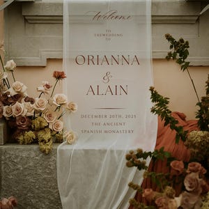May include: A sheer white wedding welcome sign with burgundy text, announcing the wedding of Orianna & Alain on December 20th, 2025, at The Ancient Spanish Monastery. The sign is draped over a stone ledge, adorned with floral arrangements.