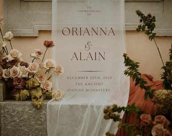 Könnte beinhalten: Ein weißes Hochzeits-Willkommensschild mit burgunderfarbenem Text, das die Hochzeit von Orianna & Alain am 20. Dezember 2025 im The Ancient Spanish Monastery ankündigt. Das Schild ist über einem Steinsims drapiert, mit Blumengestecken.