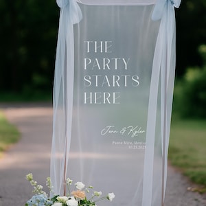 Puede incluir: Un letrero de boda transparente con el texto "THE PARTY STARTS HERE" en escritura blanca. El letrero está cubierto con tela transparente y adornado con lazos azul claro. Un arreglo floral de flores blancas, azules y melocotón se encuentra en la base.