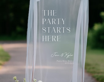 May include: A clear wedding sign with the text "THE PARTY STARTS HERE" in white script. The sign is draped with sheer fabric and adorned with light blue bows. A floral arrangement of white, blue, and peach flowers sits at the base.