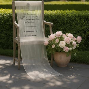 May include: A wooden chair draped with a sheer white fabric inscribed with a memorial message and a rose illustration. A terracotta pot holds a bouquet of pink roses, set on a stone patio.