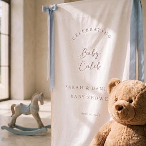 Op de afbeelding: Een verticaal gebroken wit stoffen spandoek opgehangen aan een houten frame met lichtblauwe satijnen linten aan de bovenhoeken. Het spandoek bevat donkere tekst met CELEBRATING Baby Caleb, SARAH & DANI, BABY SHOWE en April 19, 2026.