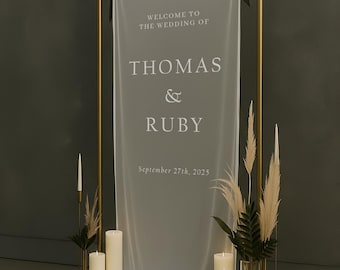 May include: A wedding welcome sign with the names THOMAS & RUBY and the date September 27th, 2025. The sign is on a gold metal frame, with a sheer white fabric. The scene includes candles and decorative plants.