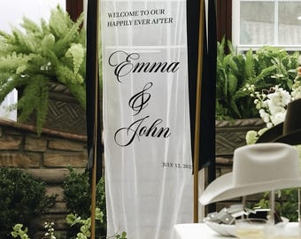 Könnte beinhalten: Ein Hochzeits-Willkommensschild mit dem Text "Welcome to our happily ever after" und den Namen "Emma & John" mit dem Datum 12. Juli 2023. Das Schild ist weiß mit schwarzen Akzenten und von Grün und Blumen umgeben.