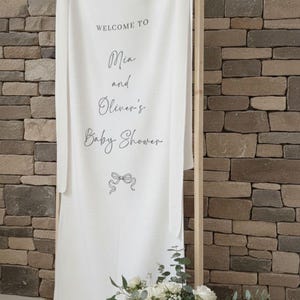 May include: A white fabric banner with the text "WELCOME TO Mia and Oliver's Baby Shower" is displayed on a wooden frame. A woven basket filled with white roses and greenery sits below the banner, on a round woven mat.