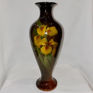 Puede incluir: Un jarrón alto de cerámica marrón con un acabado brillante. El jarrón presenta un diseño pintado a mano de flores de iris amarillas con hojas verdes. El jarrón mide aproximadamente 30 cm de alto, con una parte superior acampanada y una base redondeada.