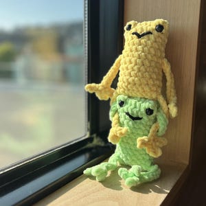 May include: Two crocheted frog plushies, one yellow and one green, stacked on top of each other. The yellow frog is on top, with its arms outstretched. The green frog has a smiling face. The plushies are on a wooden surface near a window.
