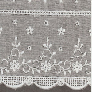 May include: Close-up of gray fabric with white floral embroidery. The design features repeating patterns of flowers, vines, and scalloped edges. Small white dots are scattered across the fabric, adding texture.
