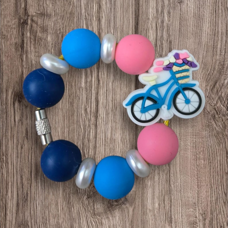 May include: A colorful bracelet featuring blue, pink, and pearl-like beads. A blue bicycle charm with a basket of flowers is attached. The bracelet is designed with a magnetic clasp.