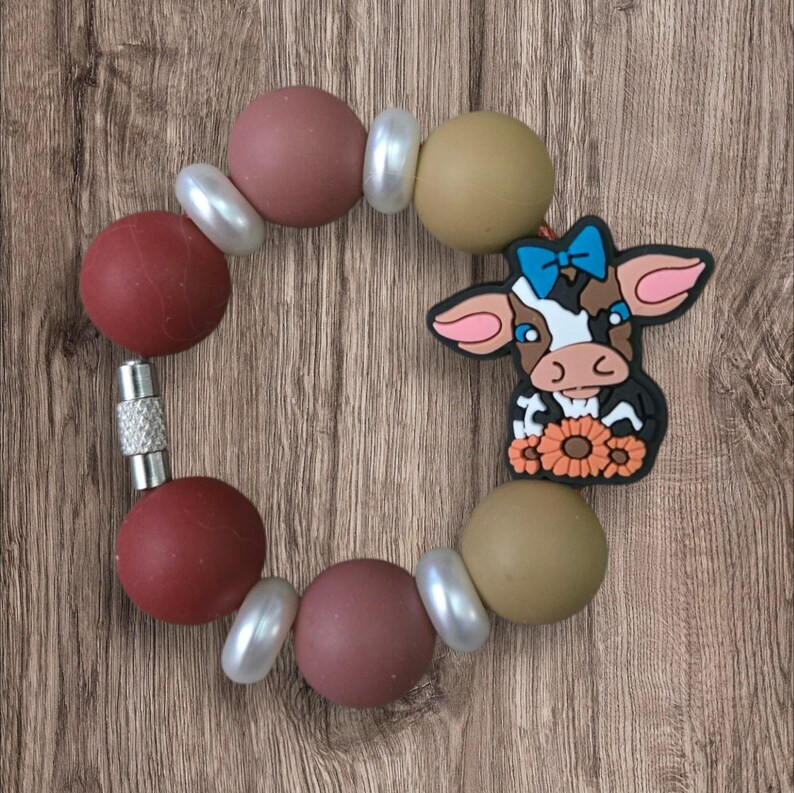 May include: A circular baby teething bracelet with a cow charm. The bracelet features large, round beads in shades of red, brown, and tan, interspersed with white pearl-like beads. The cow charm has a blue bow and a bouquet of orange flowers.