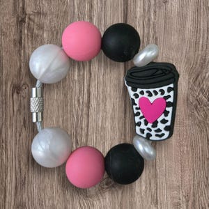 May include: A bracelet with a magnetic clasp, featuring large, round beads in pearl, pink, and black. A charm in the shape of a coffee cup with a pink heart and cow print is attached to the bracelet.