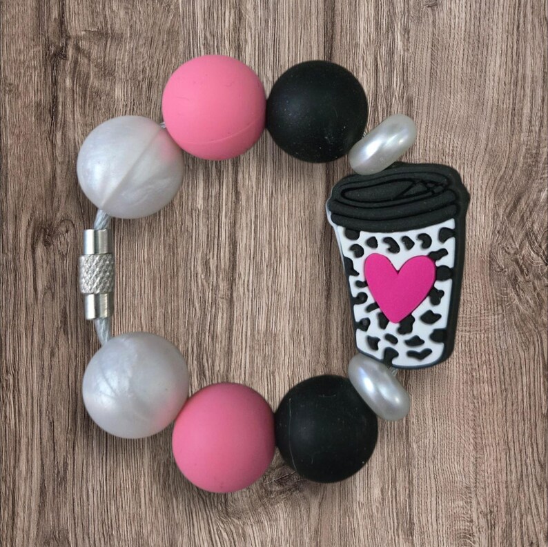 May include: A bracelet with a magnetic clasp, featuring large, round beads in pearl, pink, and black. A charm in the shape of a coffee cup with a pink heart and cow print is attached to the bracelet.