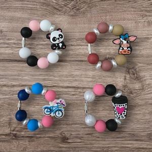 May include: Four colorful silicone bead bracelets with decorative charms. One bracelet has a panda bear charm, another has a cow, a third has a bicycle, and the last has a coffee cup with a heart.