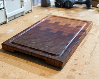 Puede incluir: Una tabla de cortar de madera grande y rectangular con un patrón de veta de madera multitono. La tabla tiene un borde elevado y dos asas en la parte inferior.