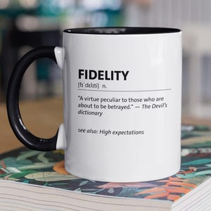 Puede incluir: Una taza de cerámica blanca con interior y asa negros. La taza presenta la definición de "FIDELITY" en texto negro, incluyendo una cita de The Devil's Dictionary. La taza está sobre un libro con un diseño floral colorido.