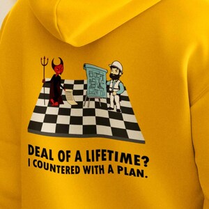 May include: Bright yellow hoodie with a graphic on the back featuring a devil and a construction worker on a checkered floor. The text reads "DEAL OF A LIFETIME? I COUNTERED WITH A PLAN."