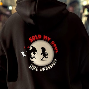 May include: Black hoodie with a circular graphic on the back. The graphic shows a running figure and the text "SOLD MY SOUL STILL UNDERPAID" in red and white. The hoodie is worn by a person walking in a city.
