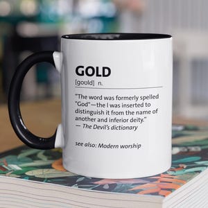 Könnte beinhalten: Weißer Keramikbecher mit schwarzem Henkel und Rand, mit dem Wort "GOLD" und einer Definition aus "The Devil's dictionary". Der Becher steht auf einem Buch mit floralem Muster.