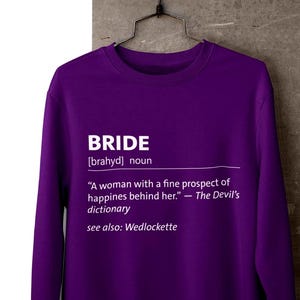 May include: A purple crewneck sweatshirt with the word "BRIDE" in white text. The text includes a definition and a reference to "Wedlockette". The sweatshirt is hanging on a black hanger.