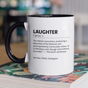 May include: White coffee mug with a black handle and rim, displaying the definition of "LAUGHTER" in black text. The mug rests on a book with a vibrant floral pattern.