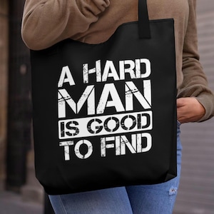 Può includere: Borsa tote nera con la scritta bianca "A Hard Man Is Good To Find". La borsa è tenuta da una persona che indossa una camicia marrone a maniche lunghe e occhiali da sole. Lo sfondo è una scena stradale sfocata.