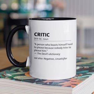 Könnte beinhalten: Weißer Keramikbecher mit schwarzem Innenraum und Henkel. Der Becher zeigt die Definition von „Critic“ aus dem Wörterbuch des Teufels. Der Becher steht auf einem Buch mit farbenfrohem Einband.