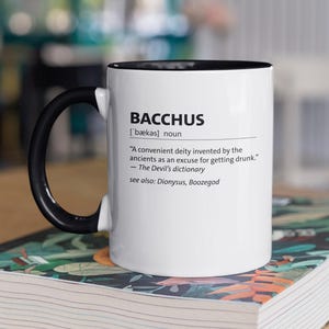 Könnte beinhalten: Weißer Keramikbecher mit schwarzem Henkel und Rand, mit der Definition von "Bacchus" aus dem Devil's Dictionary. Der Becher steht auf einem Buch mit farbenfrohem Blumendesign.