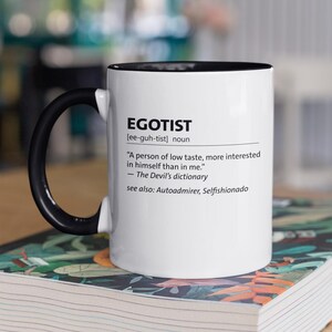Puede incluir: Taza de cerámica blanca con asa y borde negros, con la definición de "EGOTIST" en texto negro. La taza está sobre un libro con una cubierta floral colorida, colocado sobre una superficie de madera.
