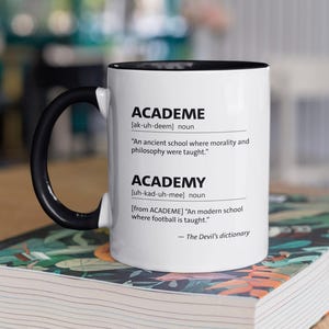 May include: A white ceramic mug with a black handle and rim, displaying the words "ACADEME" and "ACADEMY" with definitions. The mug is placed on a book with a patterned cover, against a blurred background.