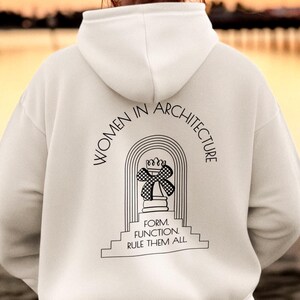 Könnte beinhalten: Ein weißer Kapuzenpullover mit dem Text "WOMEN IN ARCHITECTURE" über einer stilisierten architektonischen Grafik. Darunter steht der Text "FORM FUNCTION RULE THEM ALL". Die Person trägt den Hoodie an einem Strand, mit einem Steg im Hintergrund.