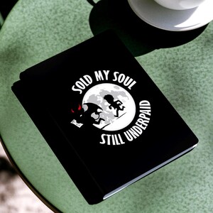May include: A black notebook with the text "SOLD MY SOUL STILL UNDERPAID" in a white circular design, with a cartoon illustration. A white coffee cup and saucer sit on a green table.