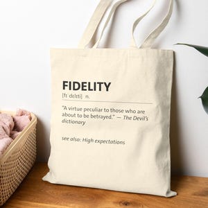 Puede incluir: Bolso tote de color crema con la palabra "FIDELITY" impresa en negro. La definición de fidelidad está impresa debajo de la palabra. La bolsa tiene dos asas y está sobre una superficie de madera.