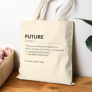 Könnte beinhalten: Eine cremefarbene Tragetasche mit dem schwarzen Aufdruck "FUTURE" und einer Definition. Die Tasche hat ein schlichtes Design und einen Griff zum Tragen. Im Hintergrund befindet sich eine rosa Decke in einem Korb.
