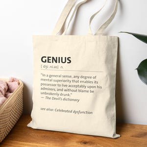 Könnte beinhalten: Cremefarbene Tragetasche mit der Definition von "GENIUS" in schwarzer Schrift. Die Definition enthält die Aussprache, ein Zitat aus The Devil's Dictionary und einen Verweis auf "Celebrated dysfunction".