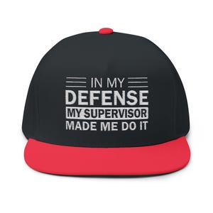 May include: Black flat-brimmed baseball cap with a red brim and button. The cap features the white text "IN MY DEFENSE MY SUPERVISOR MADE ME DO IT". A humorous accessory for casual wear.