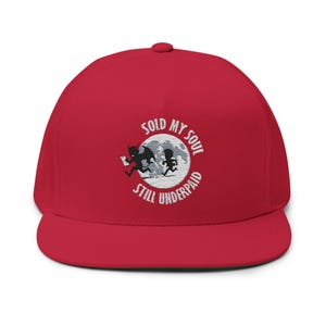 Puede incluir: Gorra de béisbol roja con visera plana. La gorra presenta un diseño bordado de un diablo persiguiendo a un hombrecito, con el texto "SOLD MY SOUL STILL UNDERPAID" en una disposición circular alrededor de una luna.