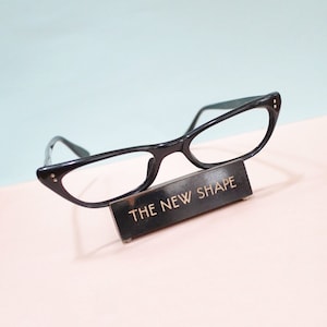 May include: Black cat-eye glasses displayed on a black rectangular stand. The stand has gold lettering that reads "THE NEW SHAPE". The glasses are set against a light blue and pink backdrop. The glasses are a classic style.