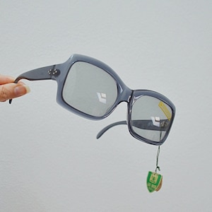 May include: Grey square-framed sunglasses with dark lenses. The sunglasses are held by a hand against a white background. A small tag is attached to the frame. The sunglasses have a retro, vintage style.